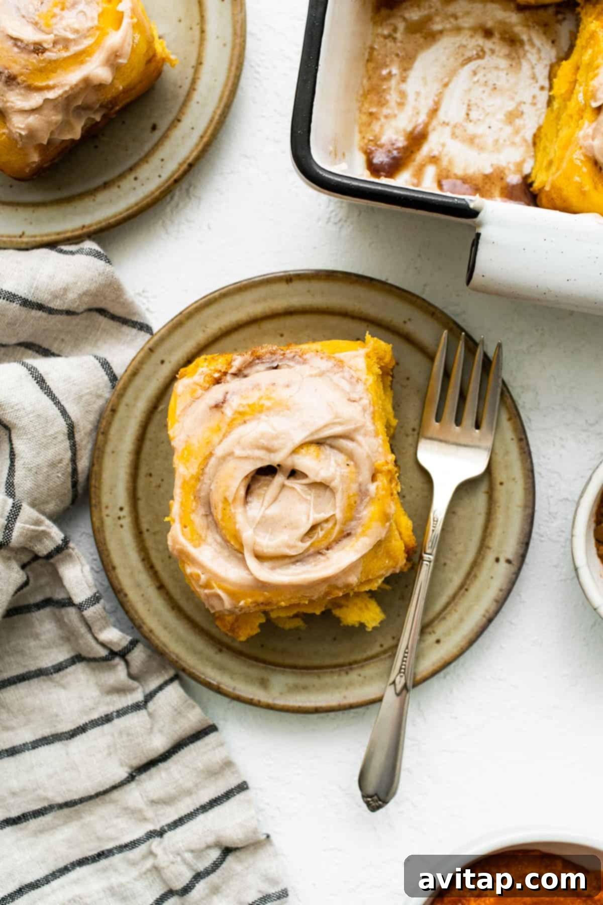 A delightful pumpkin cinnamon roll, beautifully frosted and ready to be enjoyed. A beautiful pumpkin cinnamon roll with a generous swirl of cinnamon cream cheese frosting, served on a white plate with a fork.
