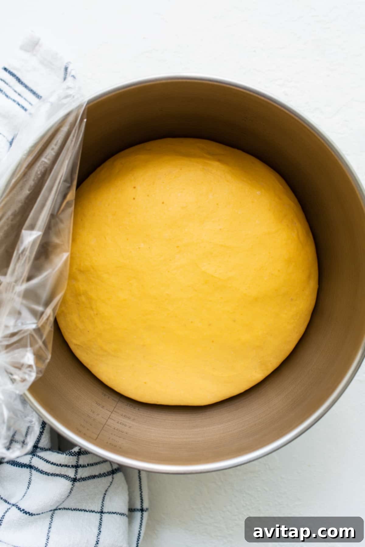 Witness the magic of a perfectly proofed pumpkin cinnamon roll dough, ready for rolling and baking into sweet treats. A perfectly risen, golden-yellow pumpkin cinnamon roll dough in a large mixing bowl, covered with plastic wrap and a tea towel.