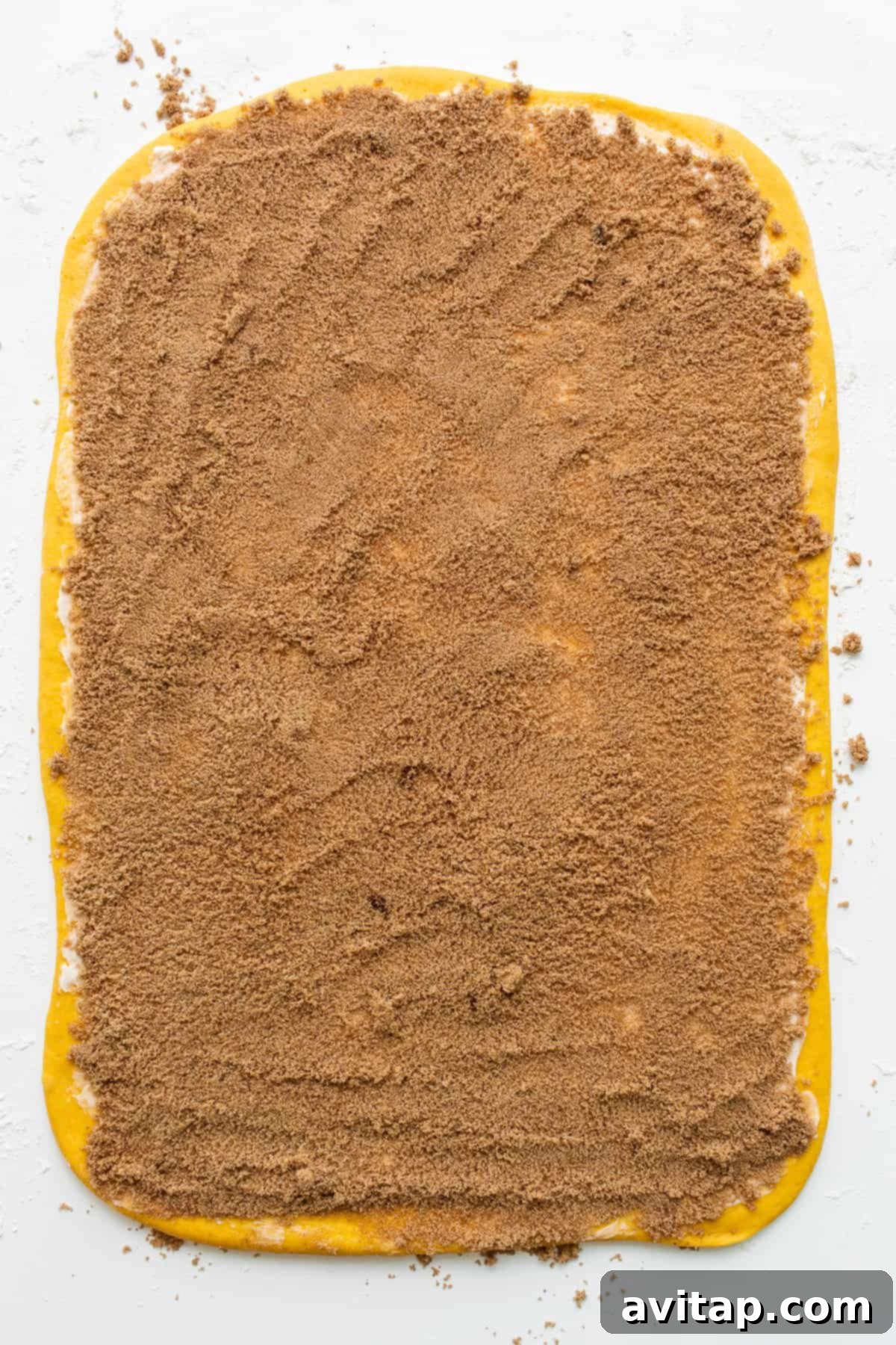 The pumpkin cinnamon roll dough is perfectly spread with the spiced brown sugar filling, creating layers of flavor before rolling. Rolled pumpkin cinnamon roll dough spread with a rich brown sugar and pumpkin pie spice filling, ready to be sliced.