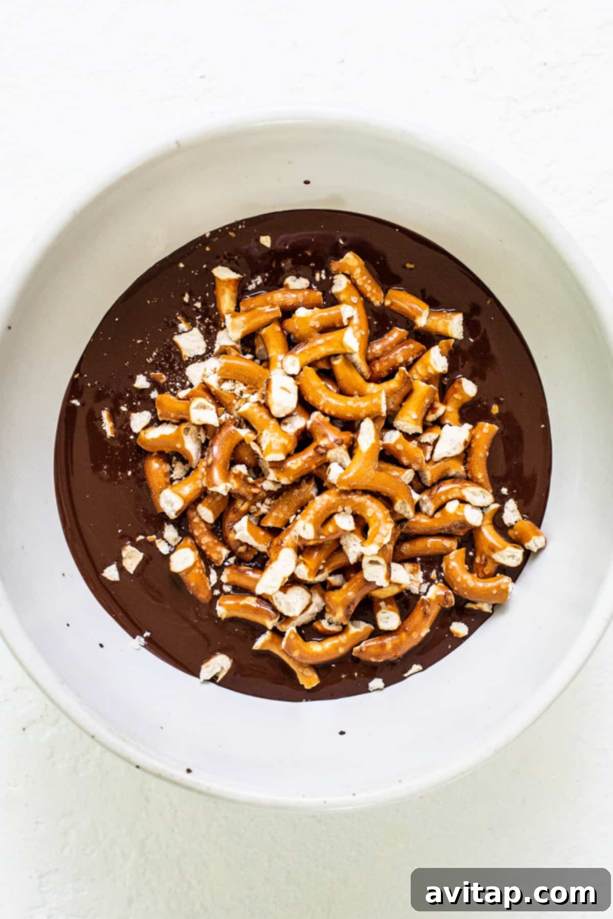 A white bowl filled with glossy melted chocolate mixed with crushed pretzels, ready to be spread over the pumpkin mousse.