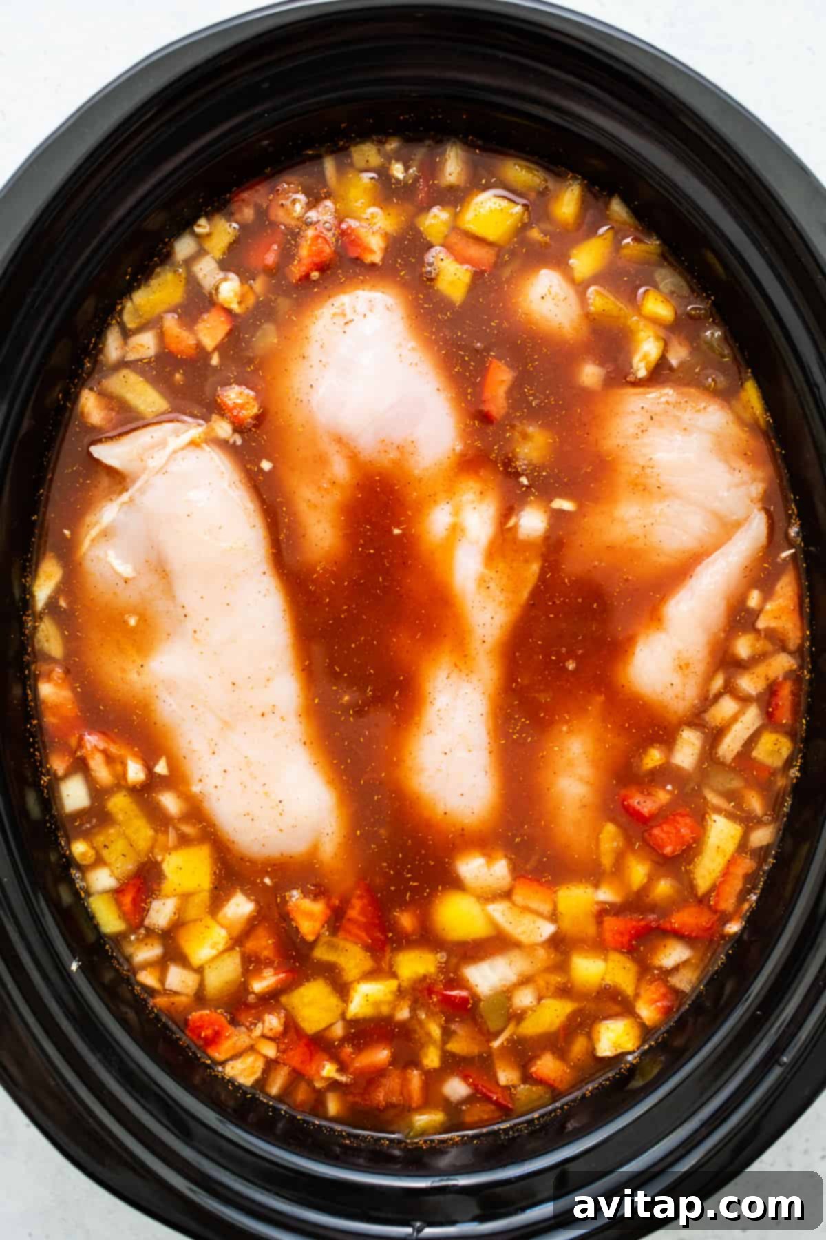Raw chicken breasts submerged among fresh vegetables and a rich tomato base in a slow cooker, illustrating the initial stage of the soup preparation.