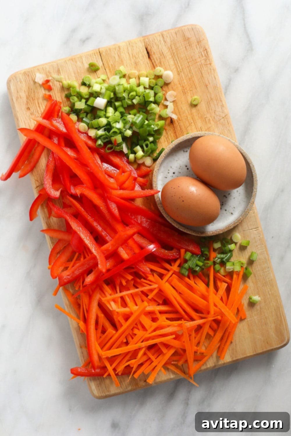 Fresh ingredients for chicken Pad Thai, including green onions, red bell pepper, eggs, and matchstick carrots, neatly arranged on a wooden cutting board.