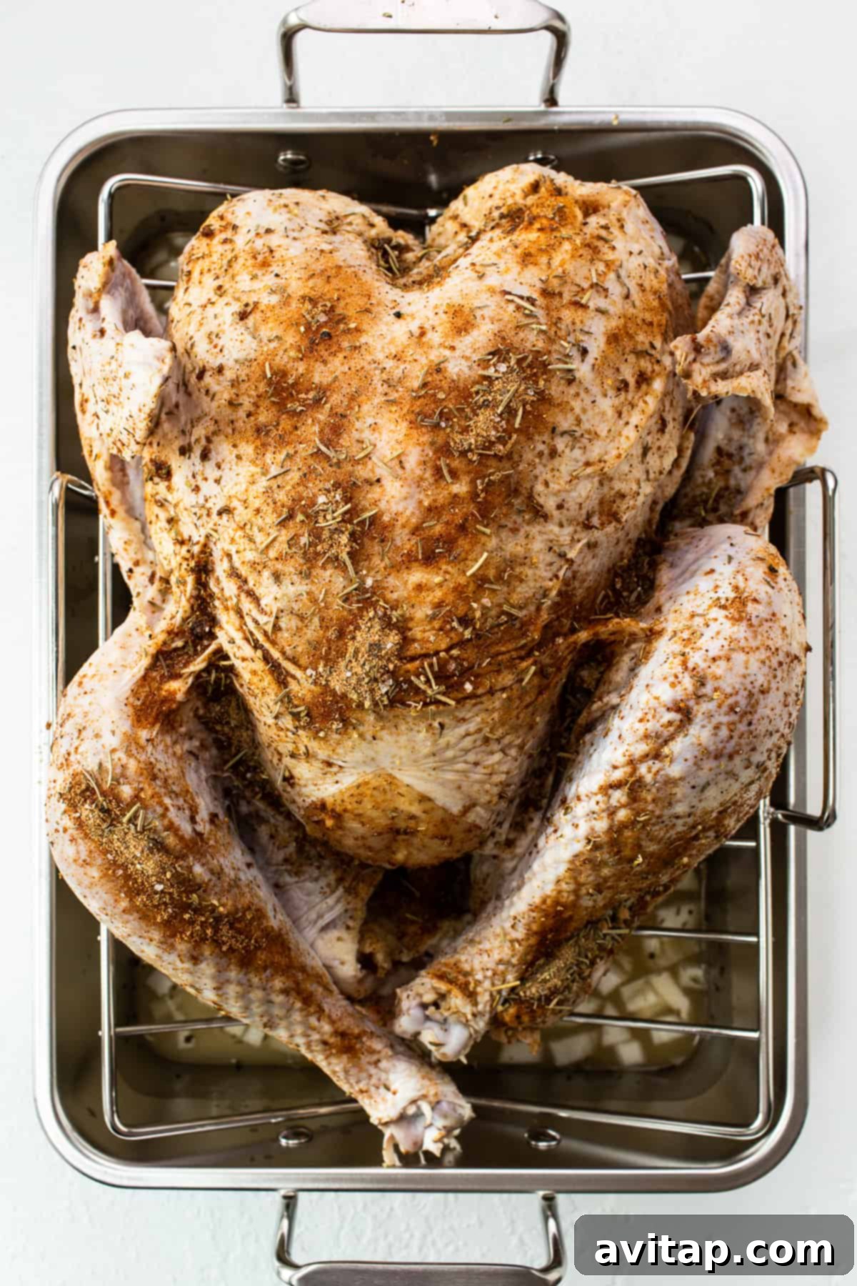 A whole turkey generously coated with a dry rub, resting in a roasting pan before cooking.