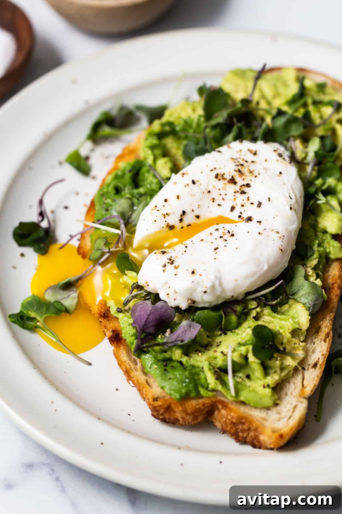 Flawless Poached Eggs Every Time 3 A perfectly poached egg resting on a slice of avocado toast, garnished with microgreens, on a white plate.