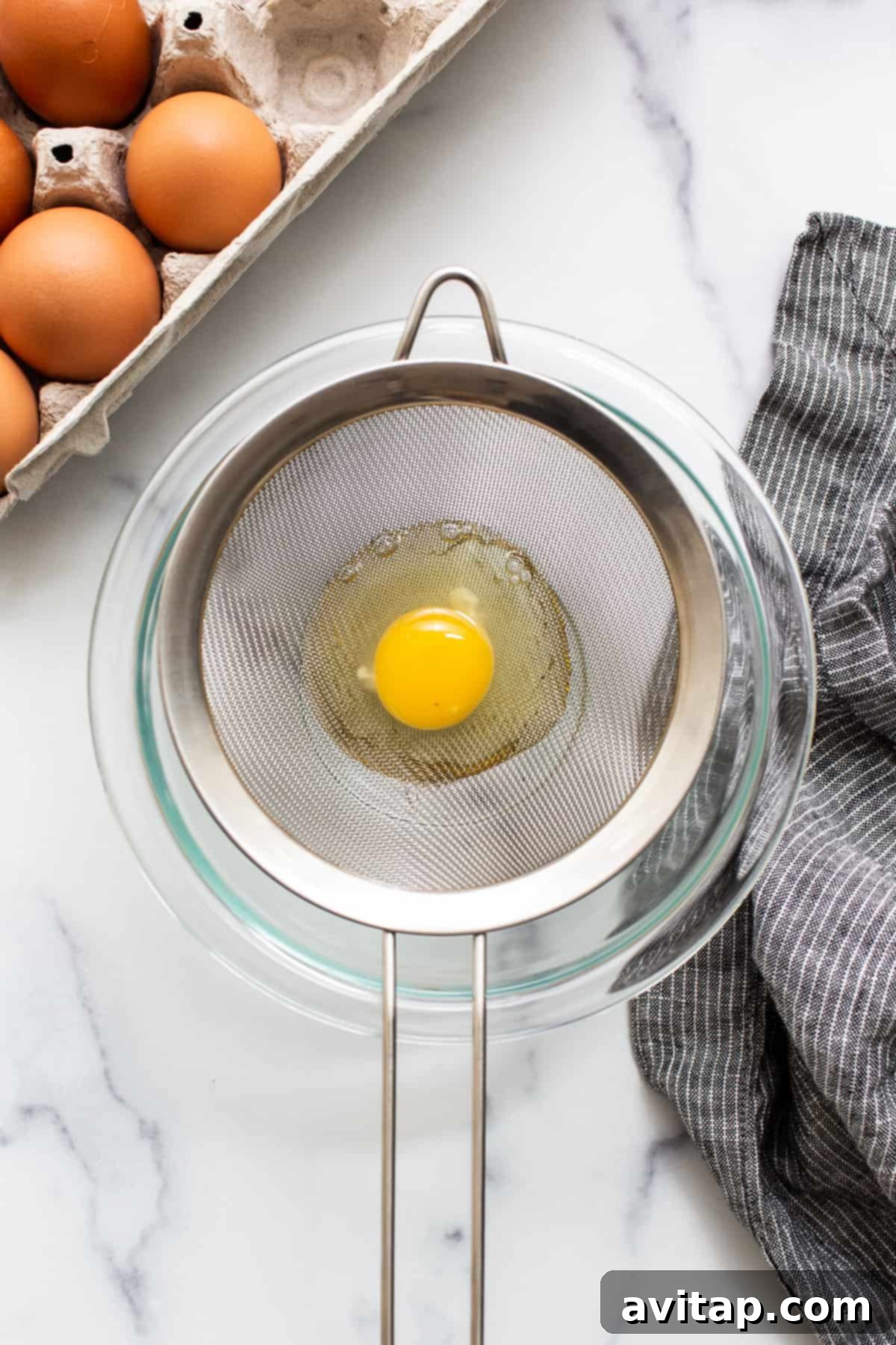 Flawless Poached Eggs Every Time 4 A raw egg being carefully strained through a fine mesh strainer over a bowl, separating the watery whites.