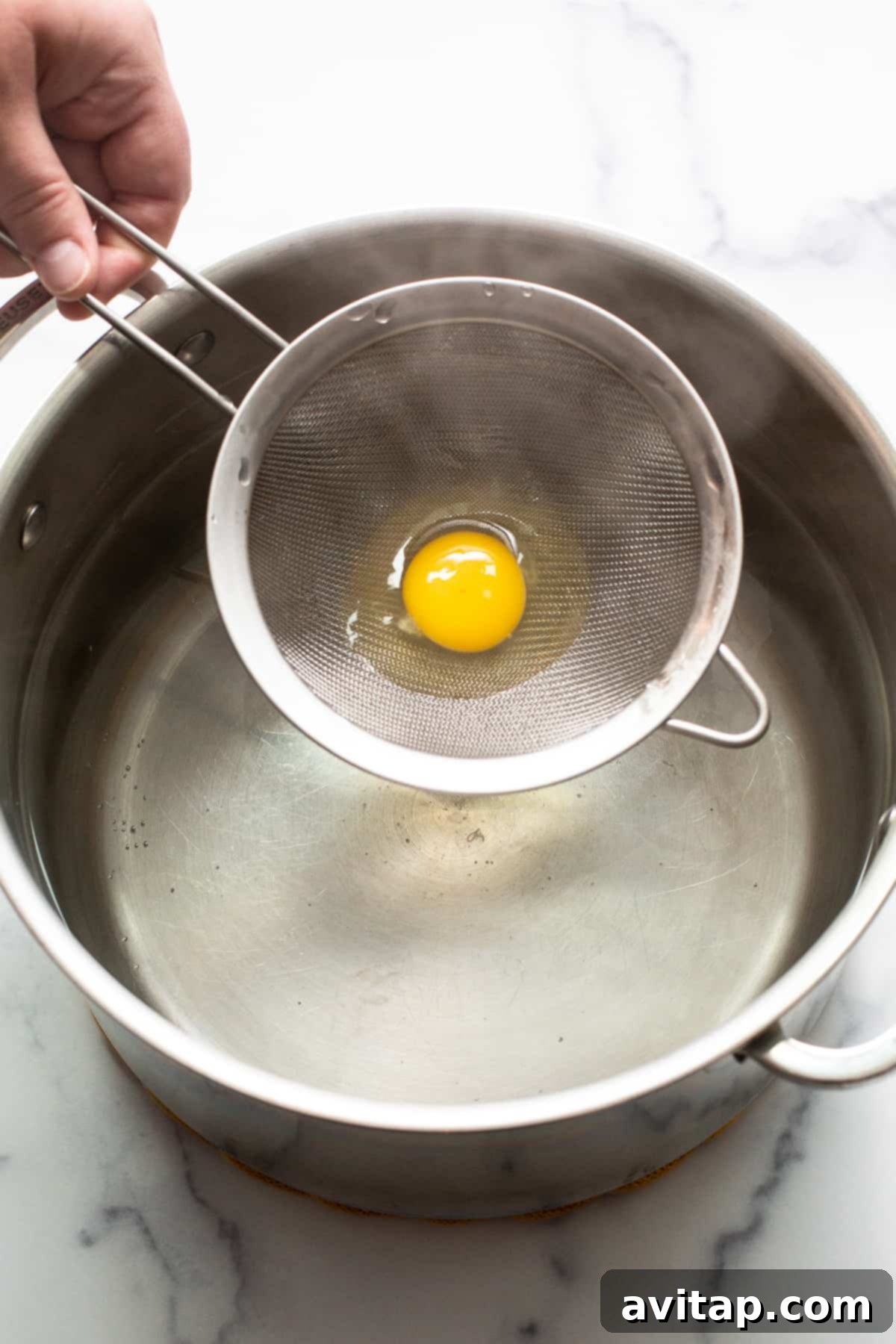 Flawless Poached Eggs Every Time 5 A raw egg, after being strained, is gently lowered in a mesh strainer into a pot of simmering water.