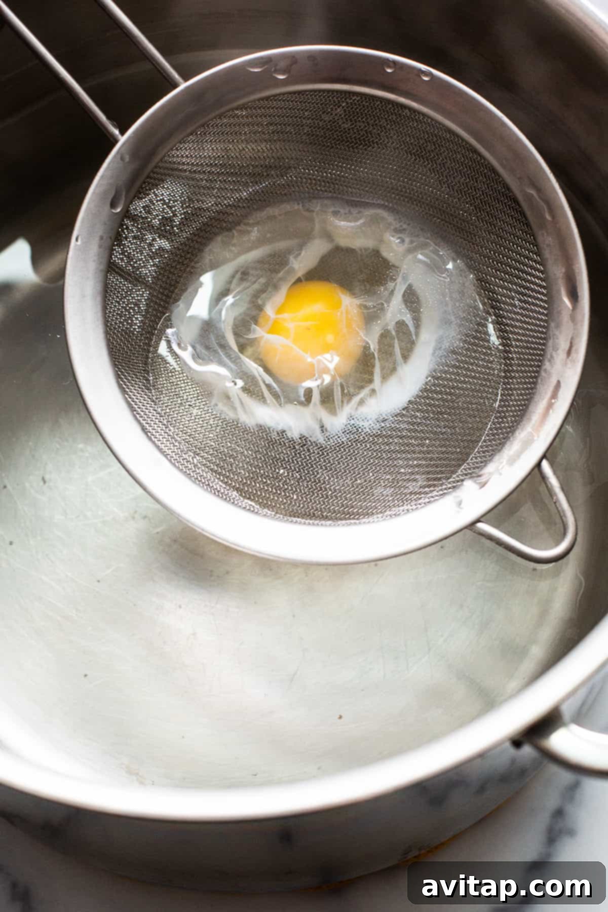Flawless Poached Eggs Every Time 6 A person carefully lowering a raw egg, held in a mesh strainer, into a pot of gently simmering water.