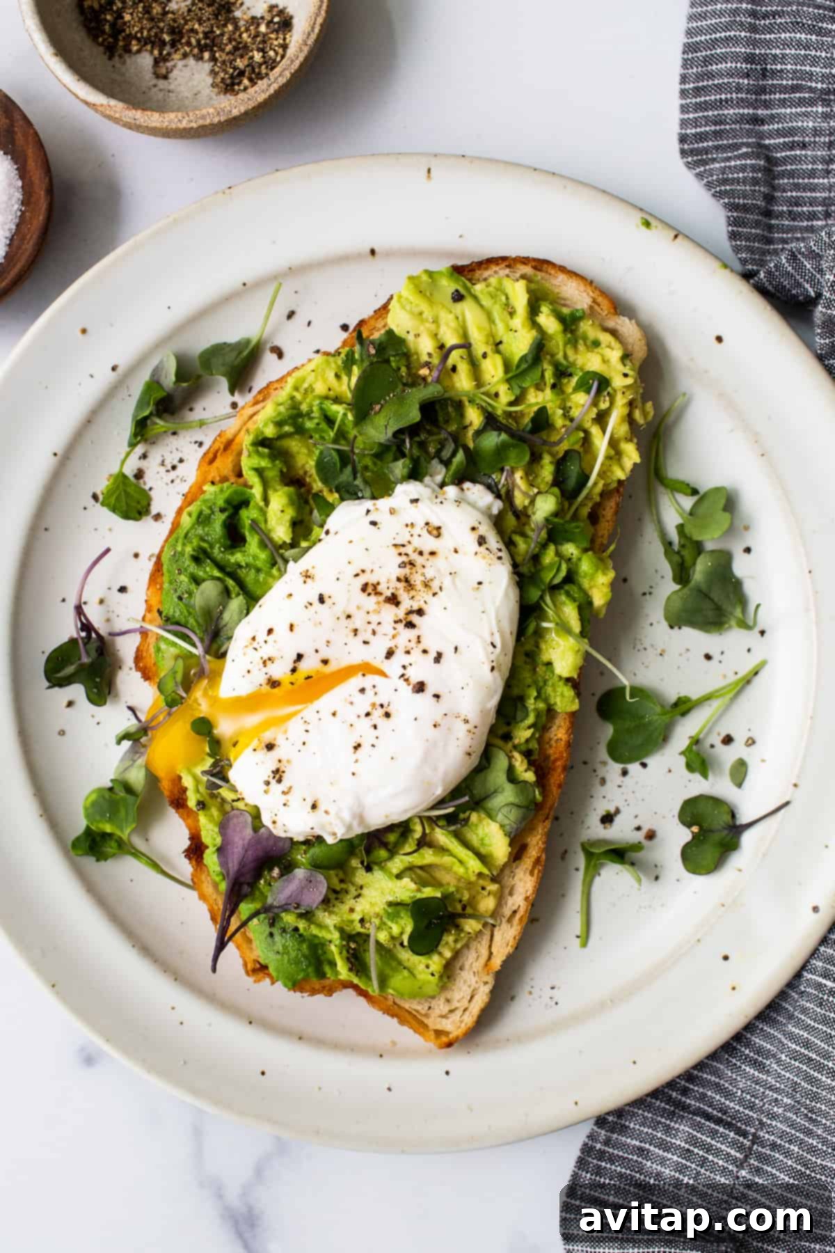 Flawless Poached Eggs Every Time 8 A perfectly poached egg on a slice of avocado toast, topped with microgreens, on a white plate with a fork.