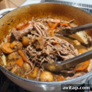 A robust pot of savory pot roast, brimming with tender, shredded beef, vibrant carrots, and soft potatoes in a rich, flavorful broth, with tongs resting on the edge.
