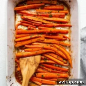 Roasted carrots in a baking dish with a wooden spoon, after initial bake and a toss, continuing to roast.