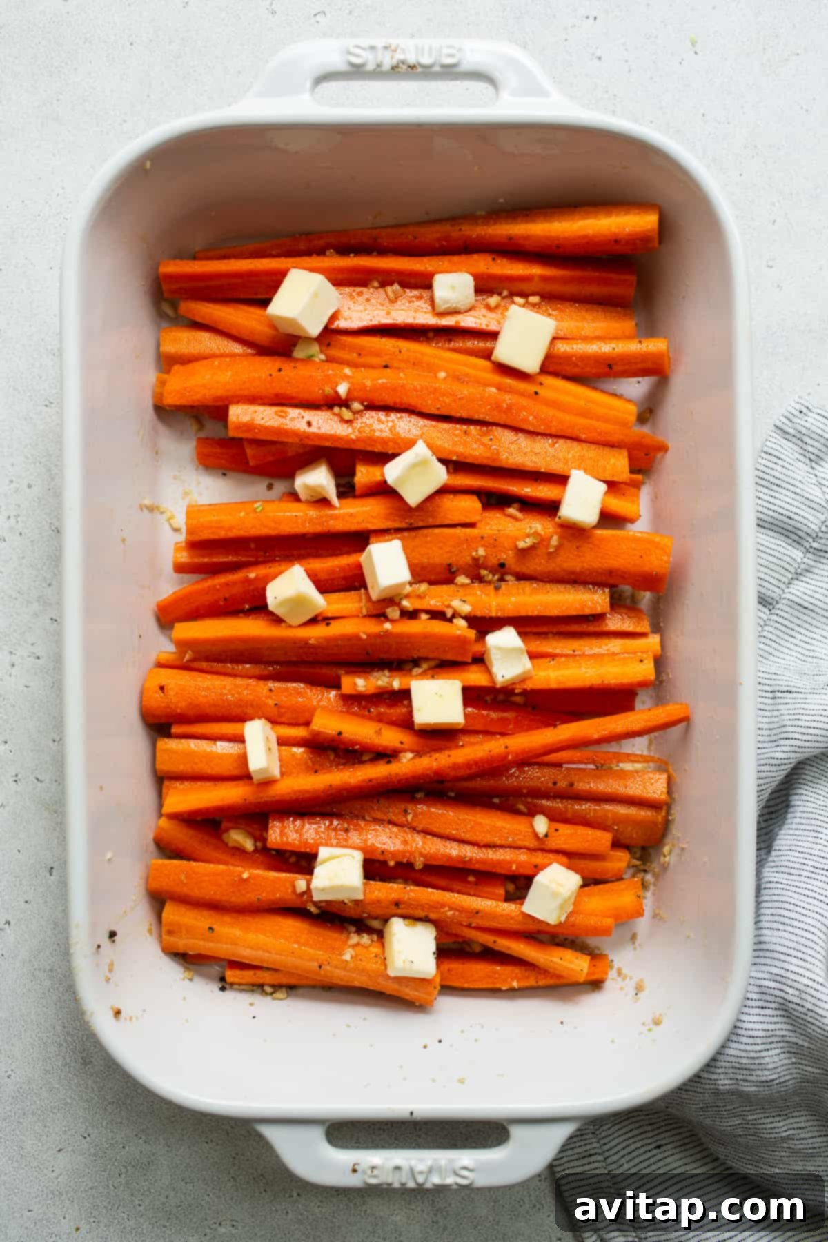 Carrots in a casserole dish, topped with cold butter chunks and seasoning, prior to roasting.