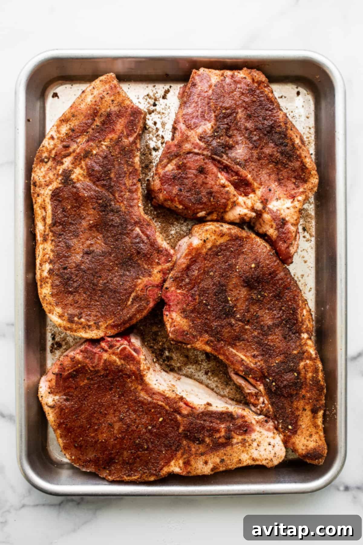 Four raw, thick-cut bone-in pork chops seasoned with a dry rub, laid out on a baking sheet, ready for air frying.
