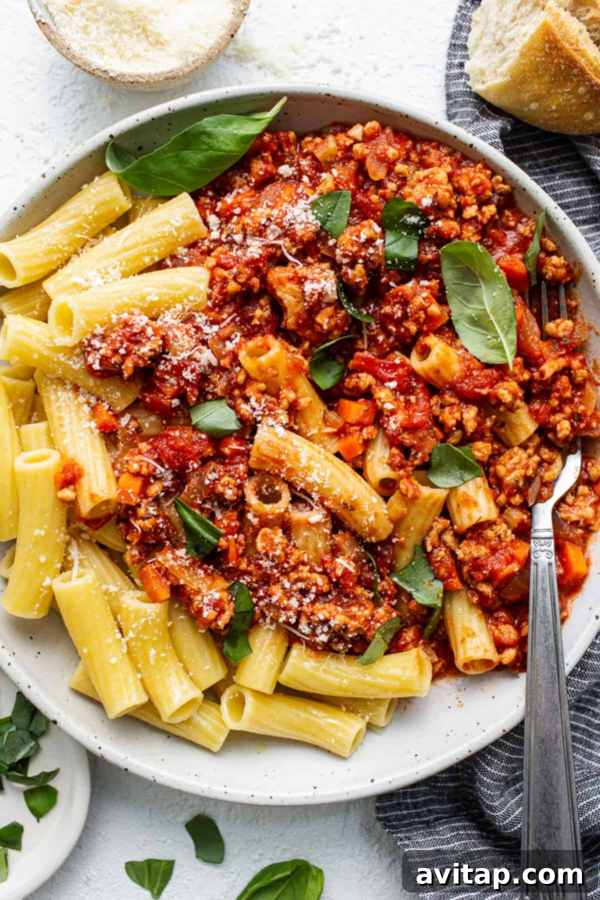 A close-up of ground chicken bolognese generously served over rotini pasta in a white bowl, garnished with fresh basil and grated Parmesan.
