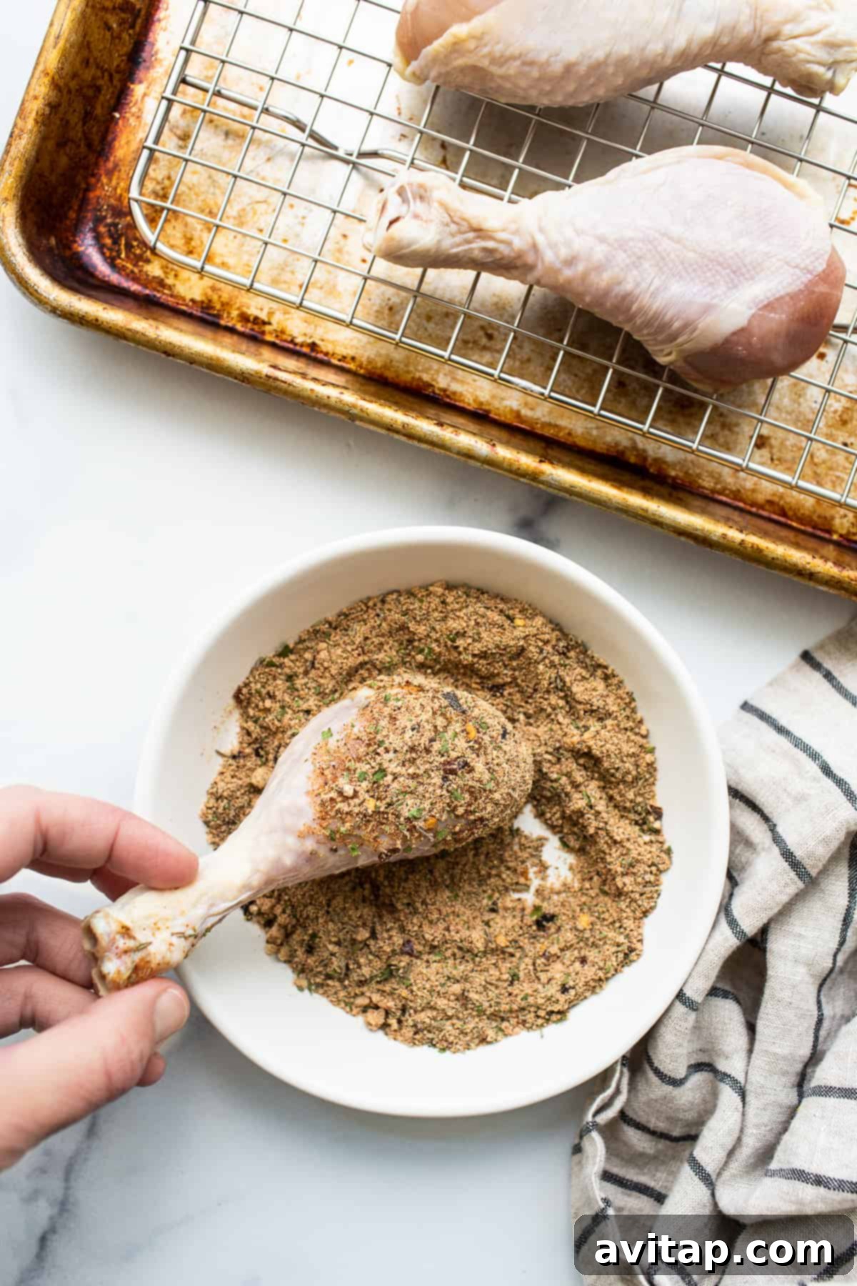 A close-up shot of raw chicken legs being generously rolled in a vibrant homemade chicken seasoning mixture on a white plate.