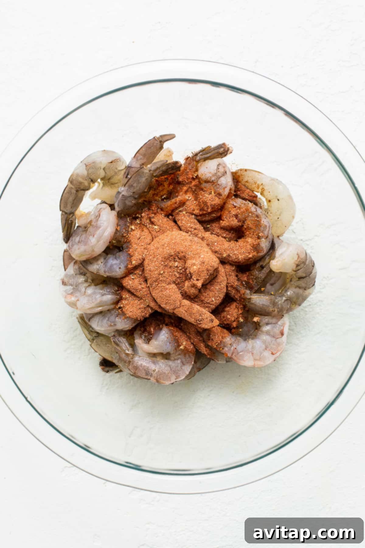 Raw jumbo shrimp ready for seasoning before broiling. Raw jumbo shrimp in a bowl, generously coated with Cajun seasoning.
