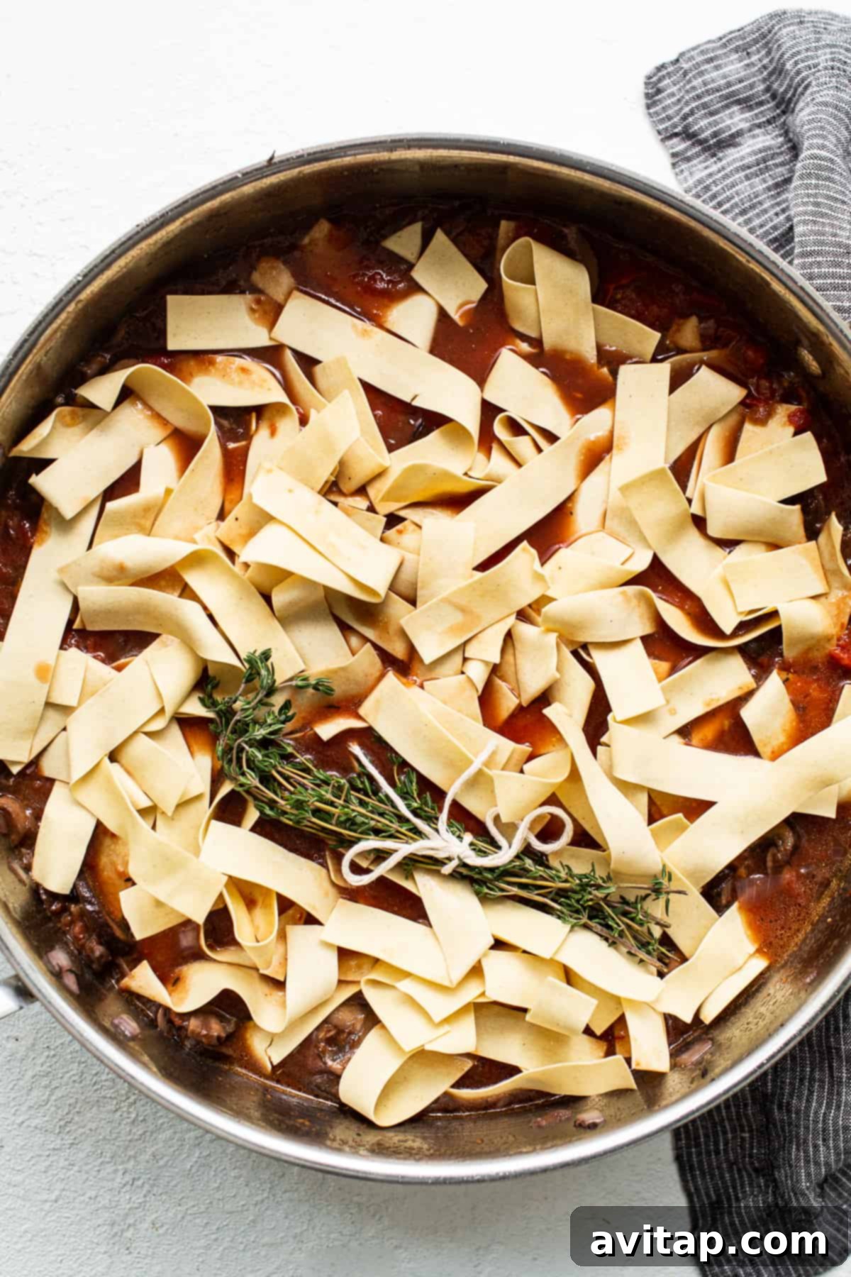 Broad pappardelle pasta being submerged into a rich, simmering red sauce in a skillet, ready to cook.
