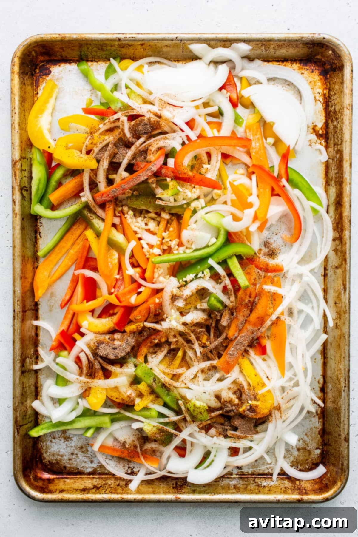 Uncooked bell peppers, onions, and garlic thinly sliced and ready for seasoning on a baking sheet.