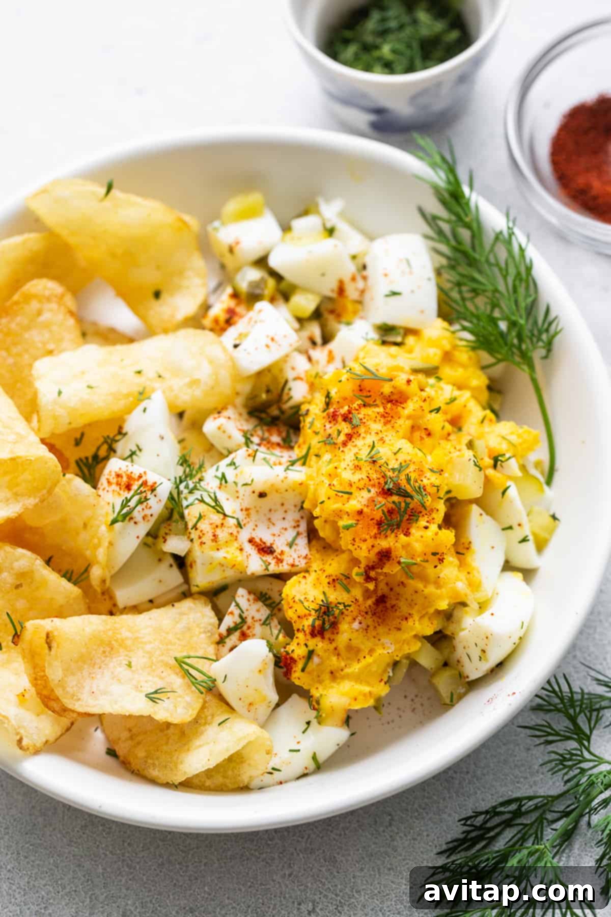 Close-up of creamy deviled egg salad in a bowl, generously garnished with vibrant fresh dill, with potato chips blurred in the background, inviting a delicious scoop.