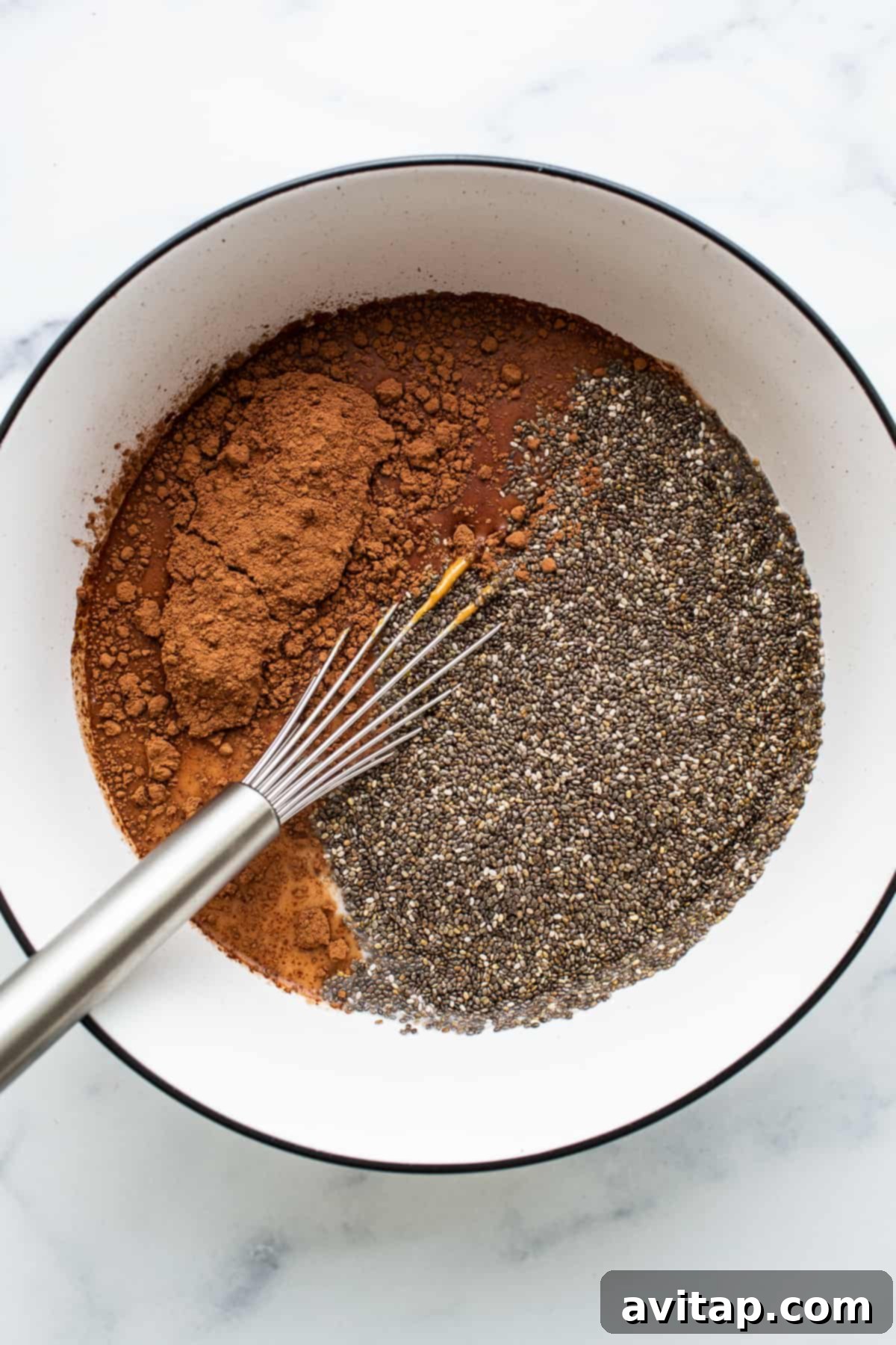 A close-up view of chia seeds and cocoa powder in a bowl, with a whisk resting beside them, illustrating the initial mixing stage of the pudding.