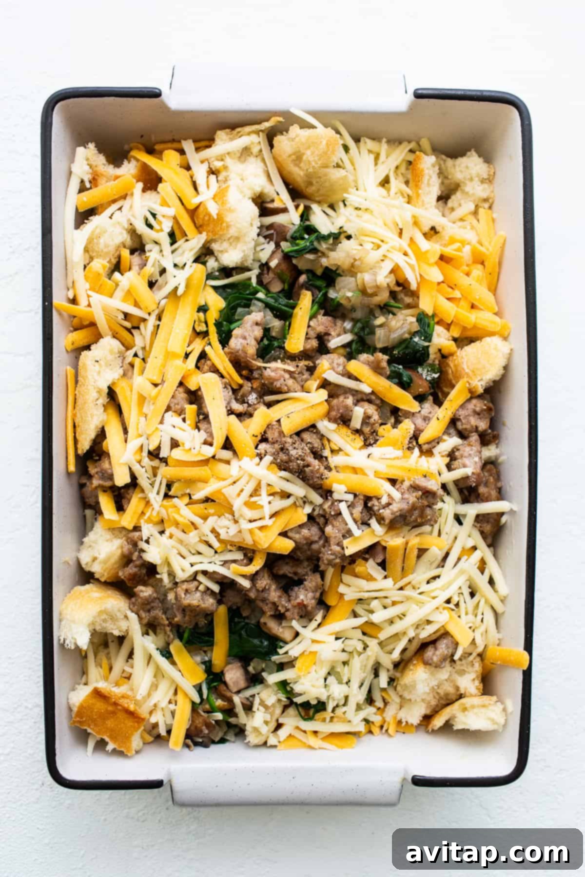 All the delicious breakfast strata ingredients beautifully layered in a casserole dish before baking, showcasing the bread, sausage, veggies, and cheese.