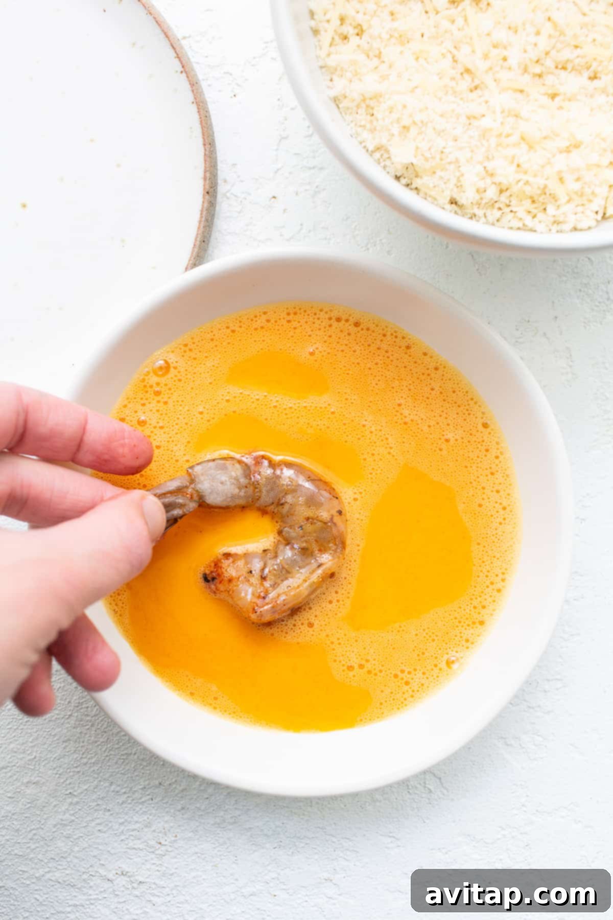 Shrimp being carefully dipped into a bowl of egg wash, ensuring full coverage.