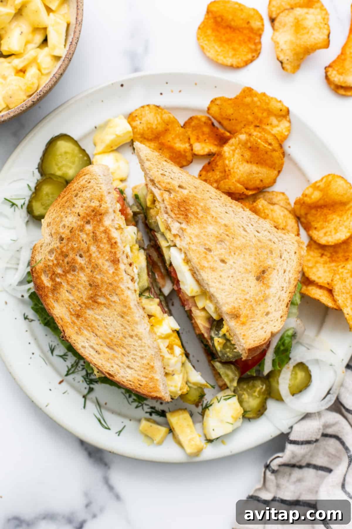 Classic Homemade Egg Salad Sandwich 7 A freshly made egg salad sandwich, cut in half, showcasing its layers of creamy filling and crisp veggies, presented on a white plate with a sprig of fresh dill.
