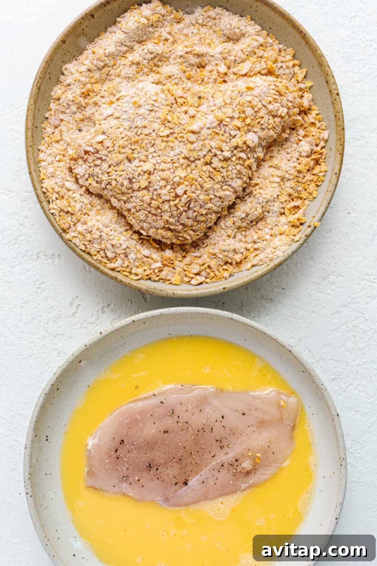 Hands dredging chicken breast in a bowl of whisked egg, preparing for coating.