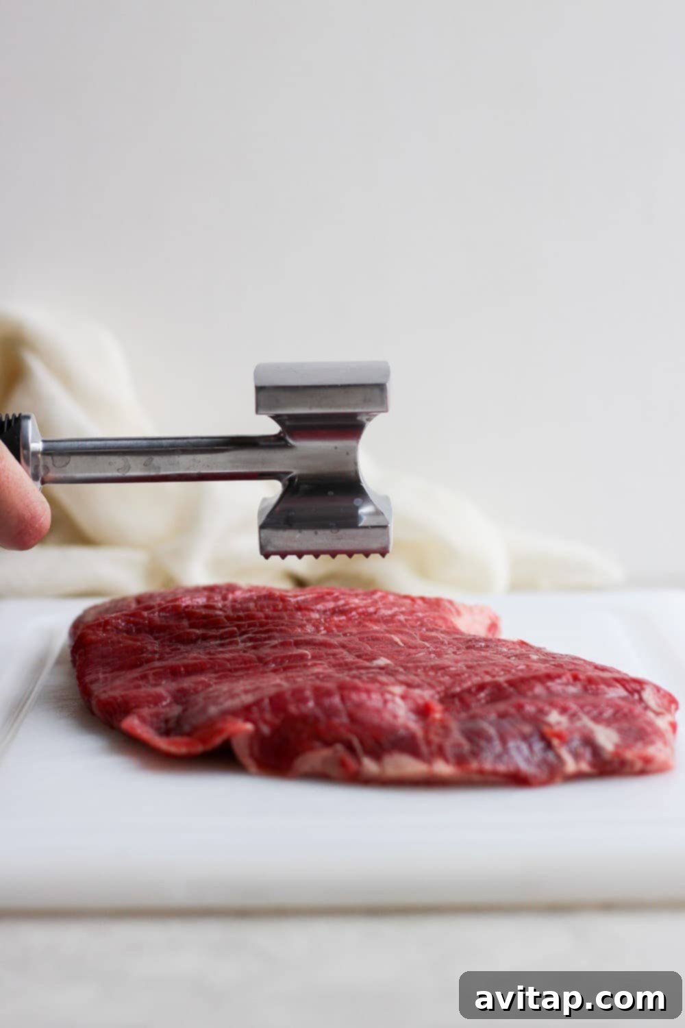A meat tenderizer actively pounding a raw flank steak on a cutting board, flattening and tenderizing the meat.
