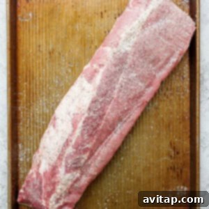 A raw rack of baby back ribs seasoned with salt and pepper on a wooden cutting board.