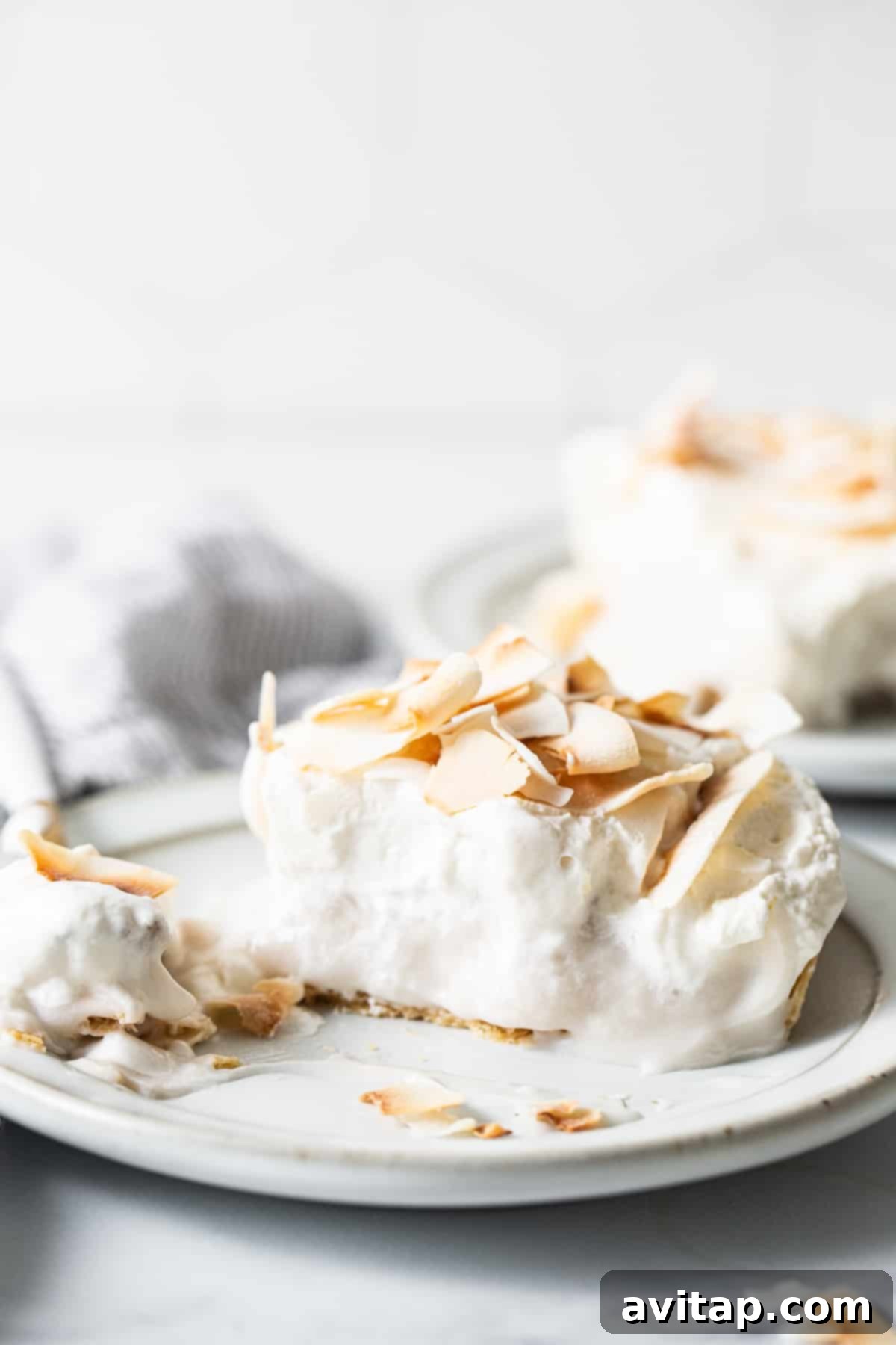 A close-up of a generous slice of coconut cream pie on a white plate, highlighting its creamy layers and toasted coconut topping.