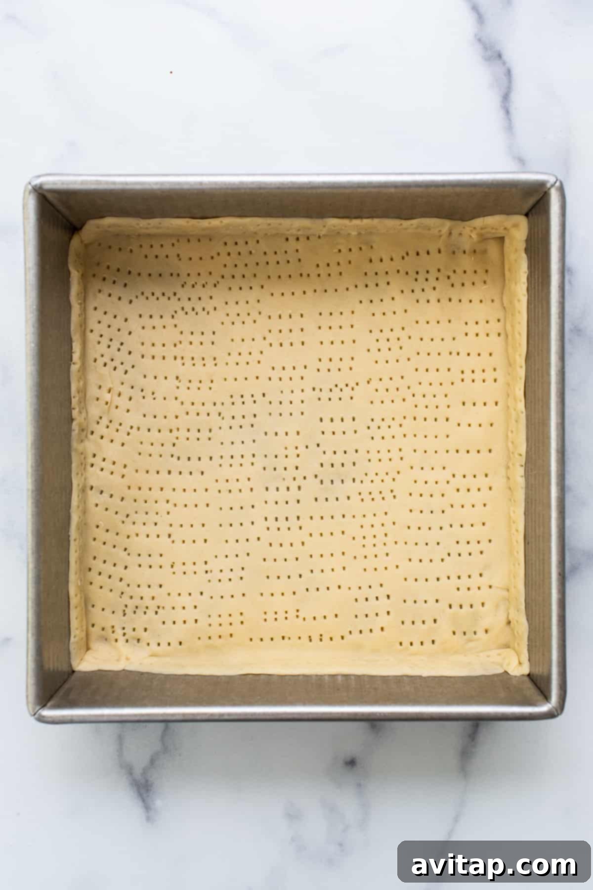 A metal baking pan with a pre-baked pie crust, showing small fork perforations, resting on a marble kitchen counter.