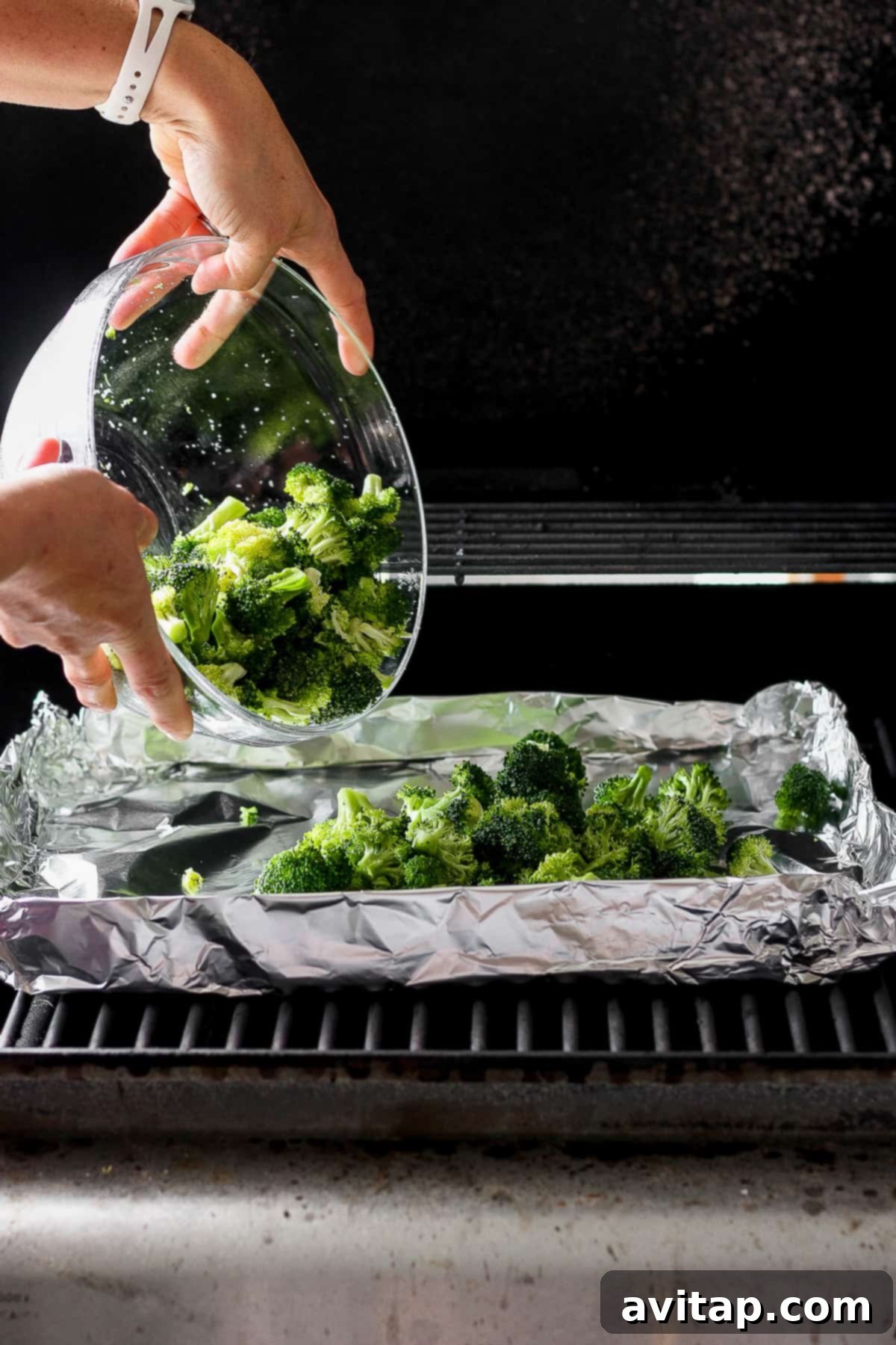 A person is putting seasoned broccoli into a large mixing bowl, preparing it for the grill.