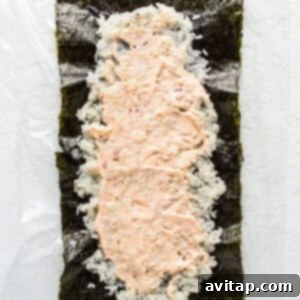 A piece of sushi being prepared on a white surface, with rice and a spread of spicy mayo.