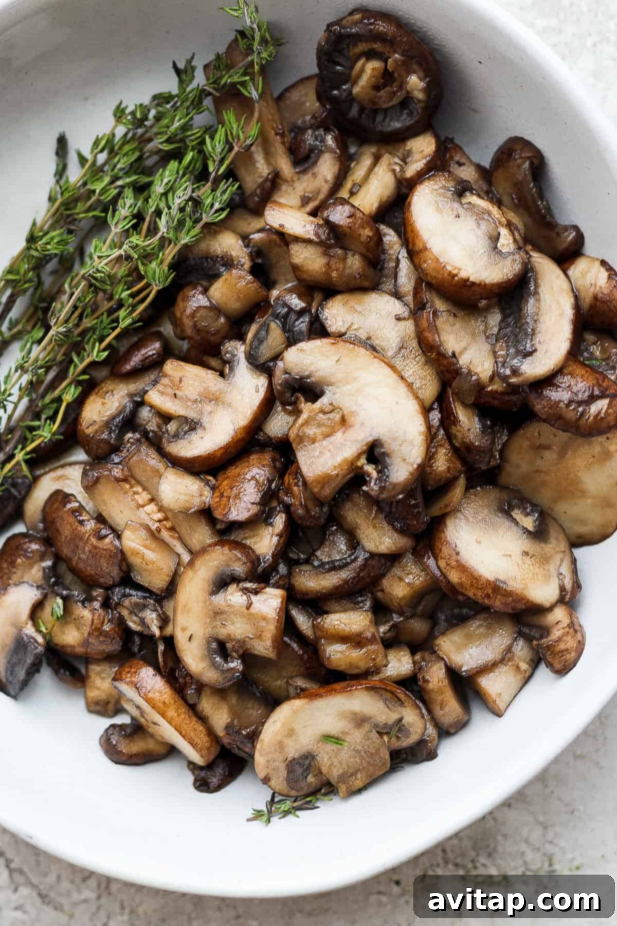 Effortless Sautéed Mushrooms 2 A white bowl filled with perfectly sautéed mushrooms, garnished with a sprig of fresh thyme, showcasing their golden-brown color and appealing texture.