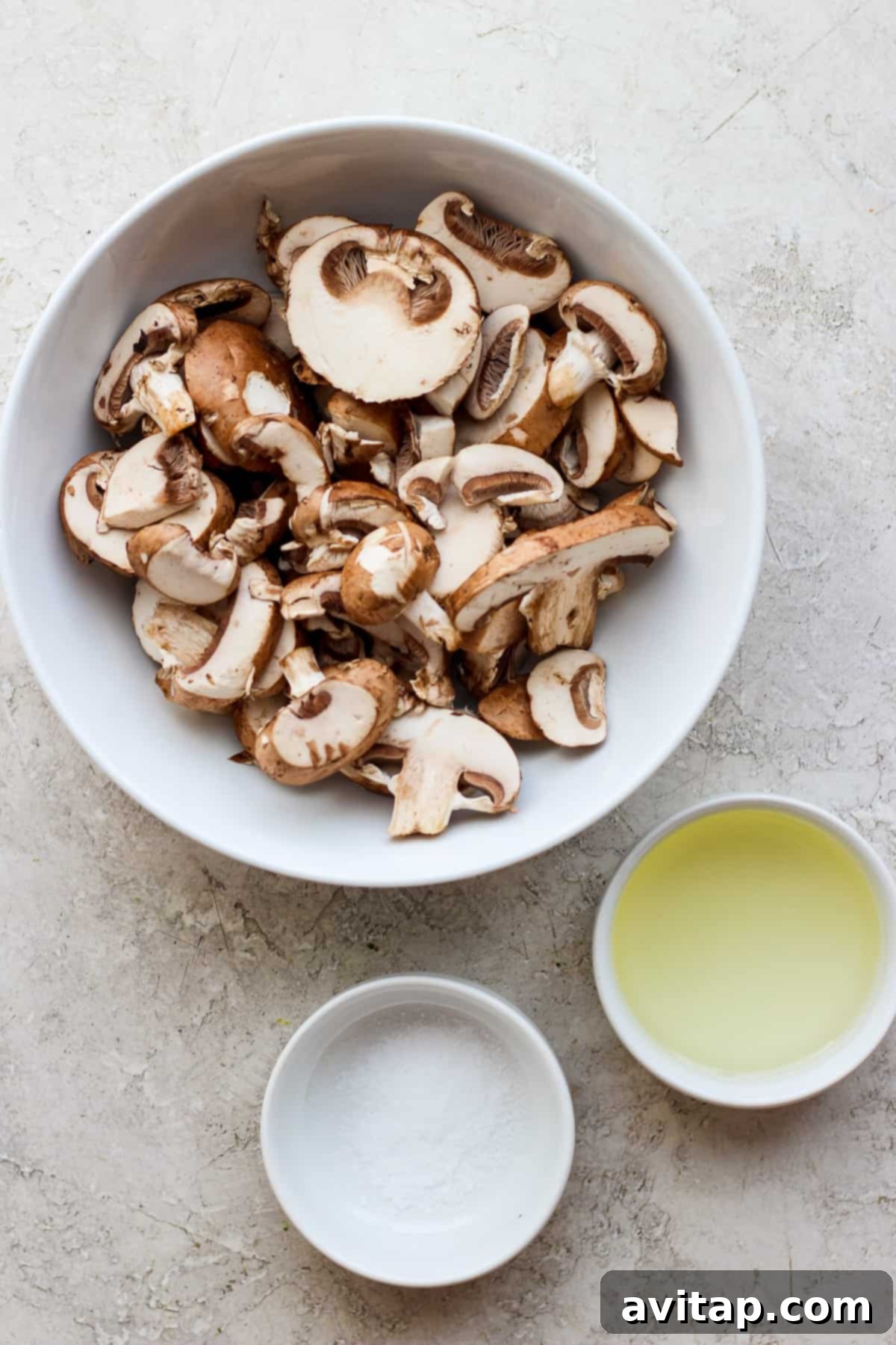 Effortless Sautéed Mushrooms 3 Fresh Bella mushrooms in a white bowl next to a smaller bowl of olive oil, ready for sautéing.