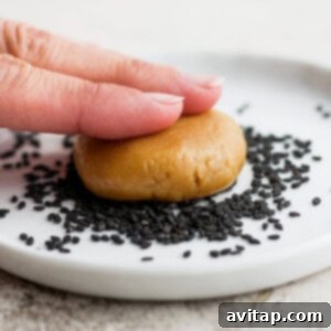 A hand holding a flattened tahini cookie dough ball, coated with sesame seeds on one side.