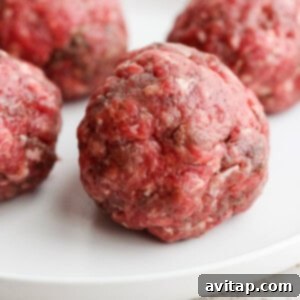 Four raw meatballs on a white plate, seasoned and ready for cooking.