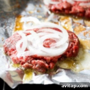 Beef burgers topped with onions cooking on aluminum foil on a grill.