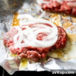 The Mushroom Swiss Burger 16 Ground beef patties with thinly sliced onions on top, cooking on aluminum foil directly on a hot grill.