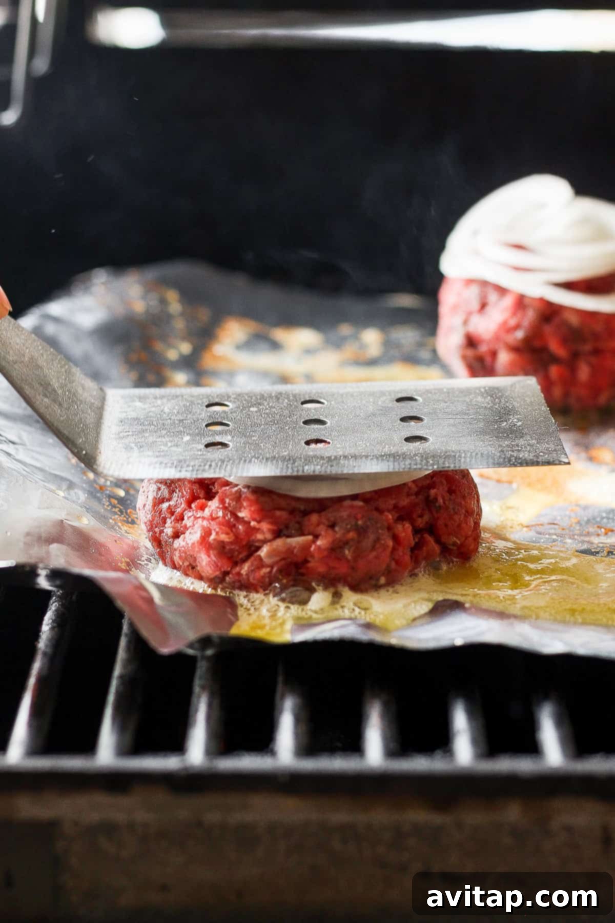 A person uses a spatula to press down on beef patties cooking on a grill, ensuring a perfect sear.