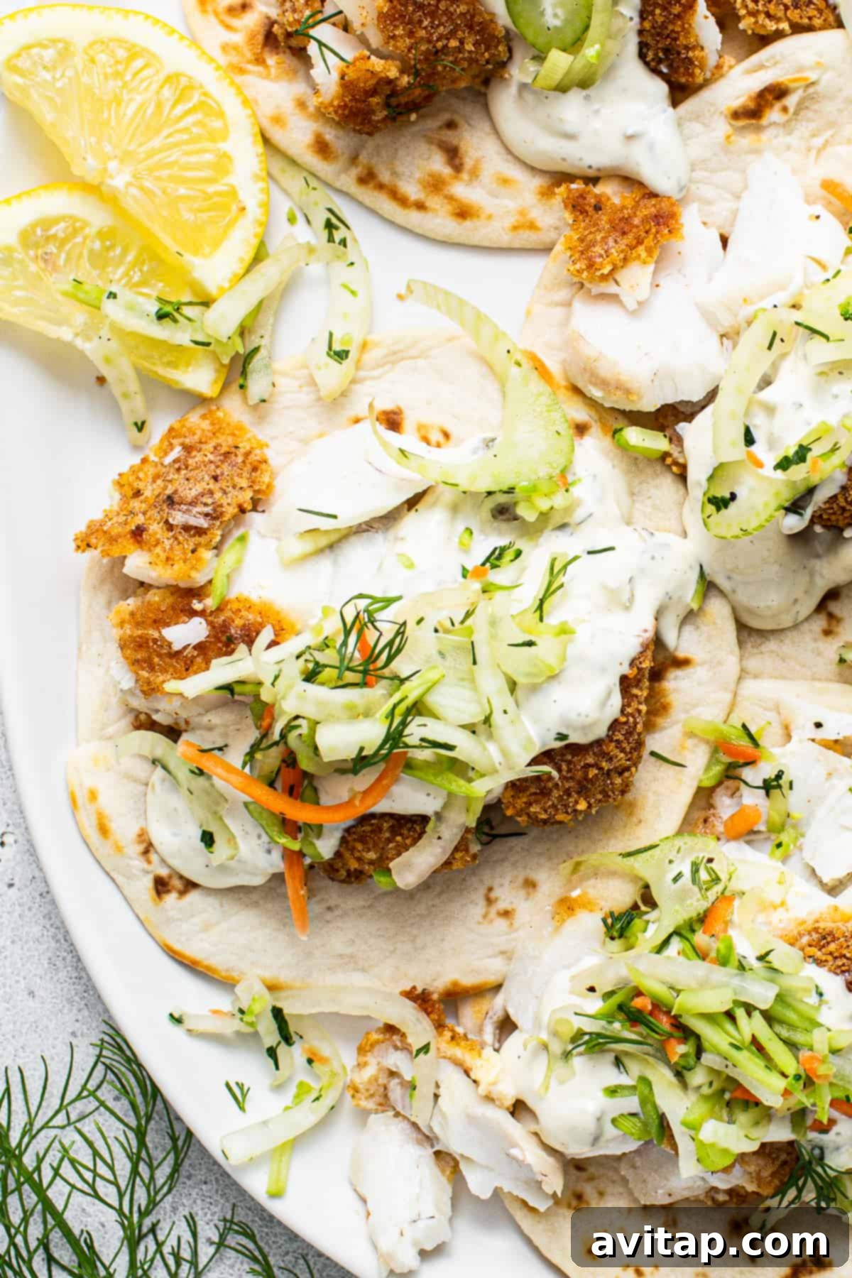 Two fish tacos generously filled with crispy fish, colorful slaw, and a drizzle of sauce, served on a plate with fresh lemon wedges.