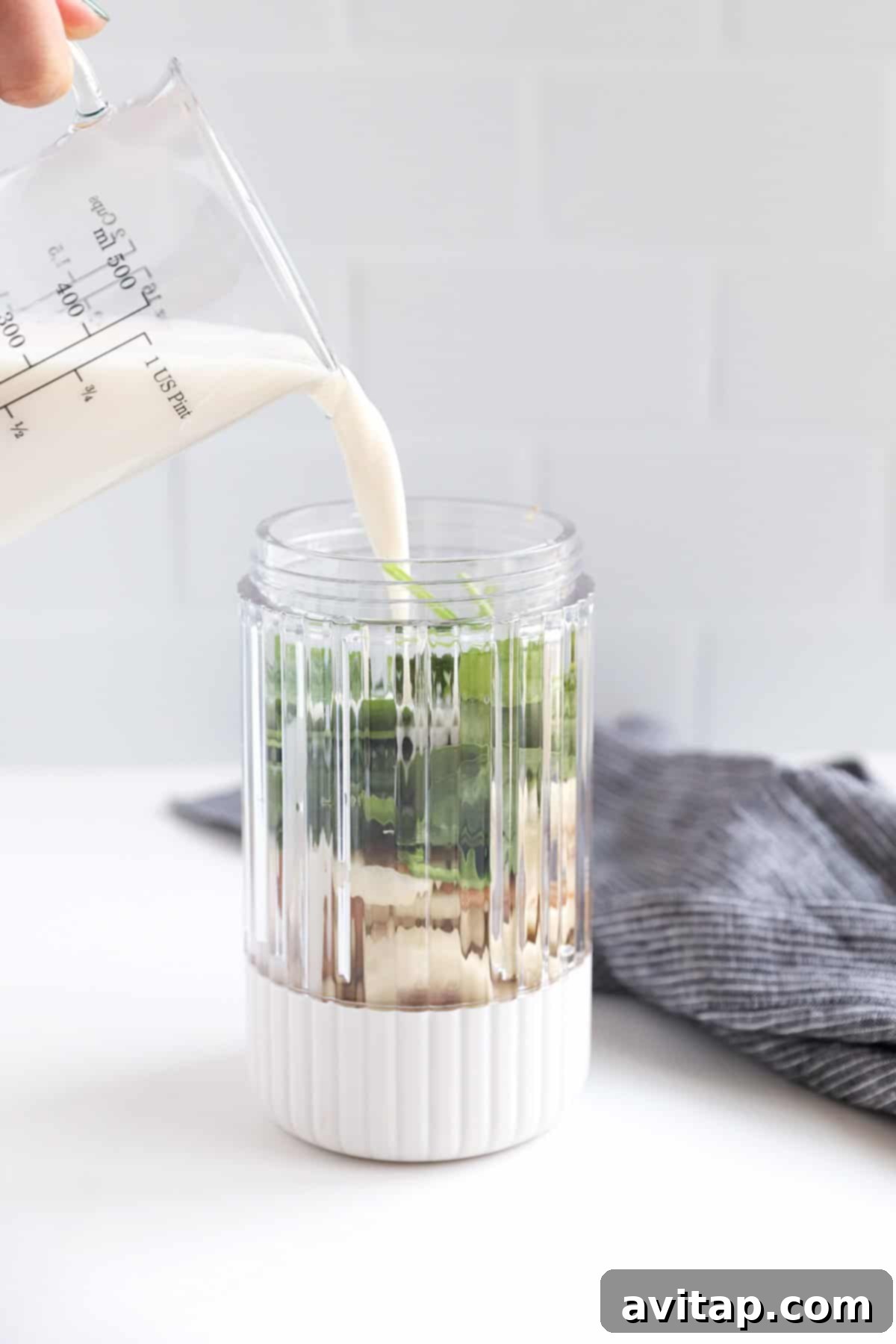 A person pouring almond milk into a glass pitcher, preparing smoothie ingredients.