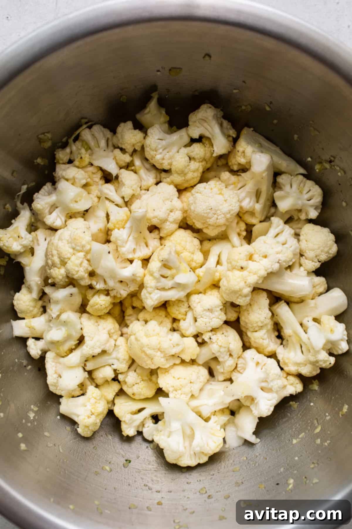 Cauliflower florets seasoned with a generous drizzle of olive oil, minced garlic, salt, and pepper in a large mixing bowl, ready to be tossed.
