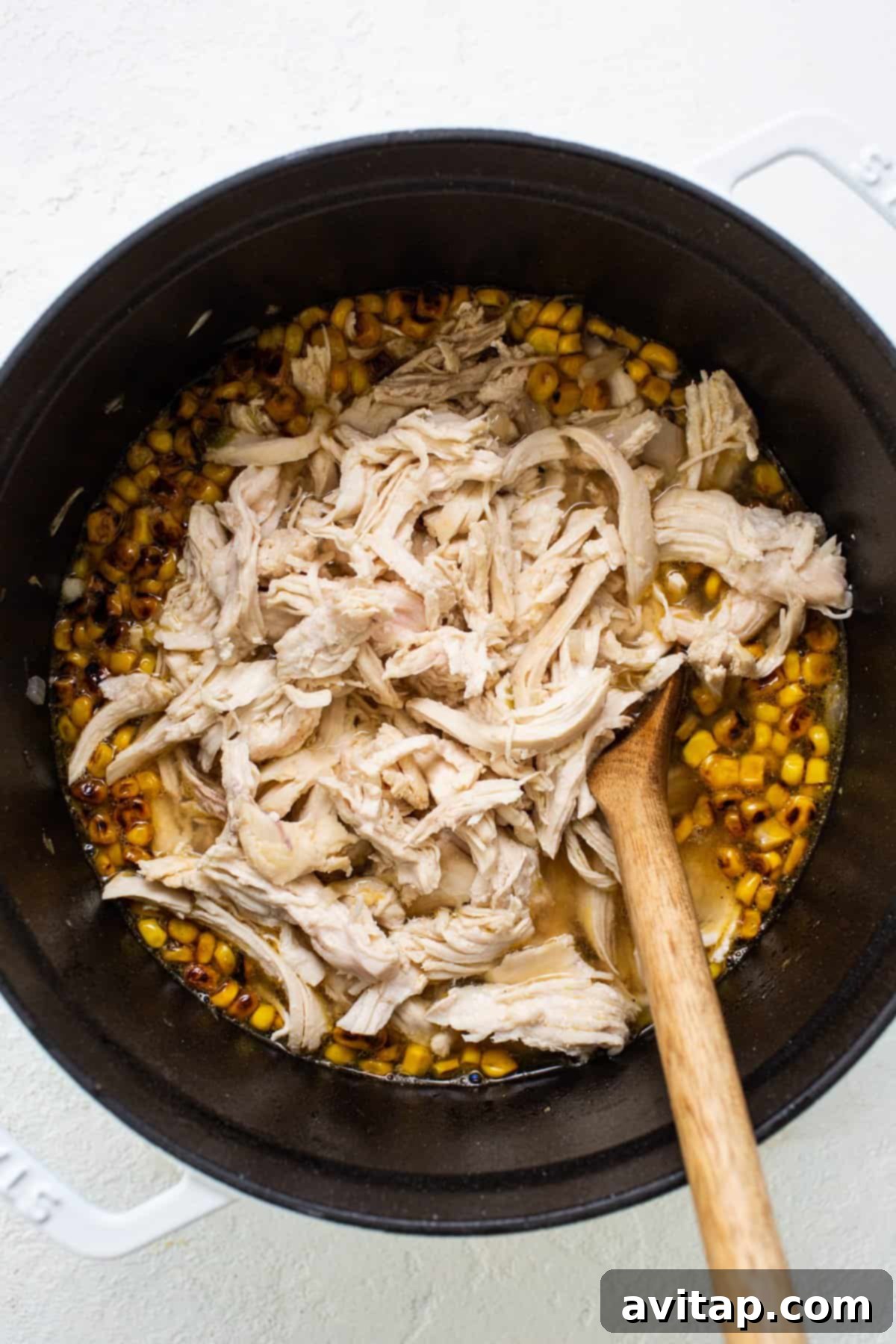 Close-up view of succulent chicken and golden corn simmering in a rich, savory broth within a large, rustic pot, with a wooden spoon resting nearby, hinting at the depth of flavors developing.