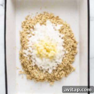 A white baking dish containing uncooked quinoa, minced onion, and garlic, ready for the next step.