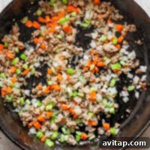 A cast iron skillet filled with cooking ground Italian sausage, onions, garlic, carrots, and celery mixture.