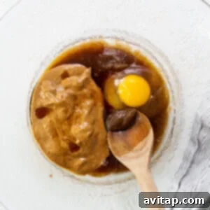 An egg being cracked into a glass bowl with other wet ingredients and a wooden spoon.