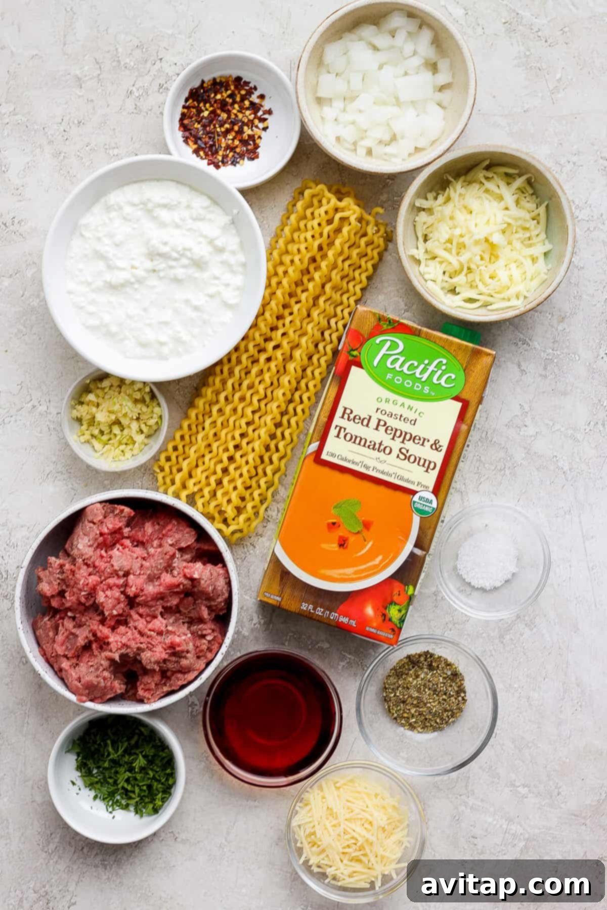 A selection of fresh ingredients including ground beef, creamy tomato soup, various cheeses, and pasta noodles laid out on a kitchen counter, ready for cooking.