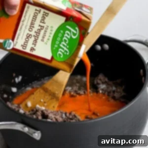 A hand pouring creamy tomato soup into a large pot with cooked ground beef and aromatics.