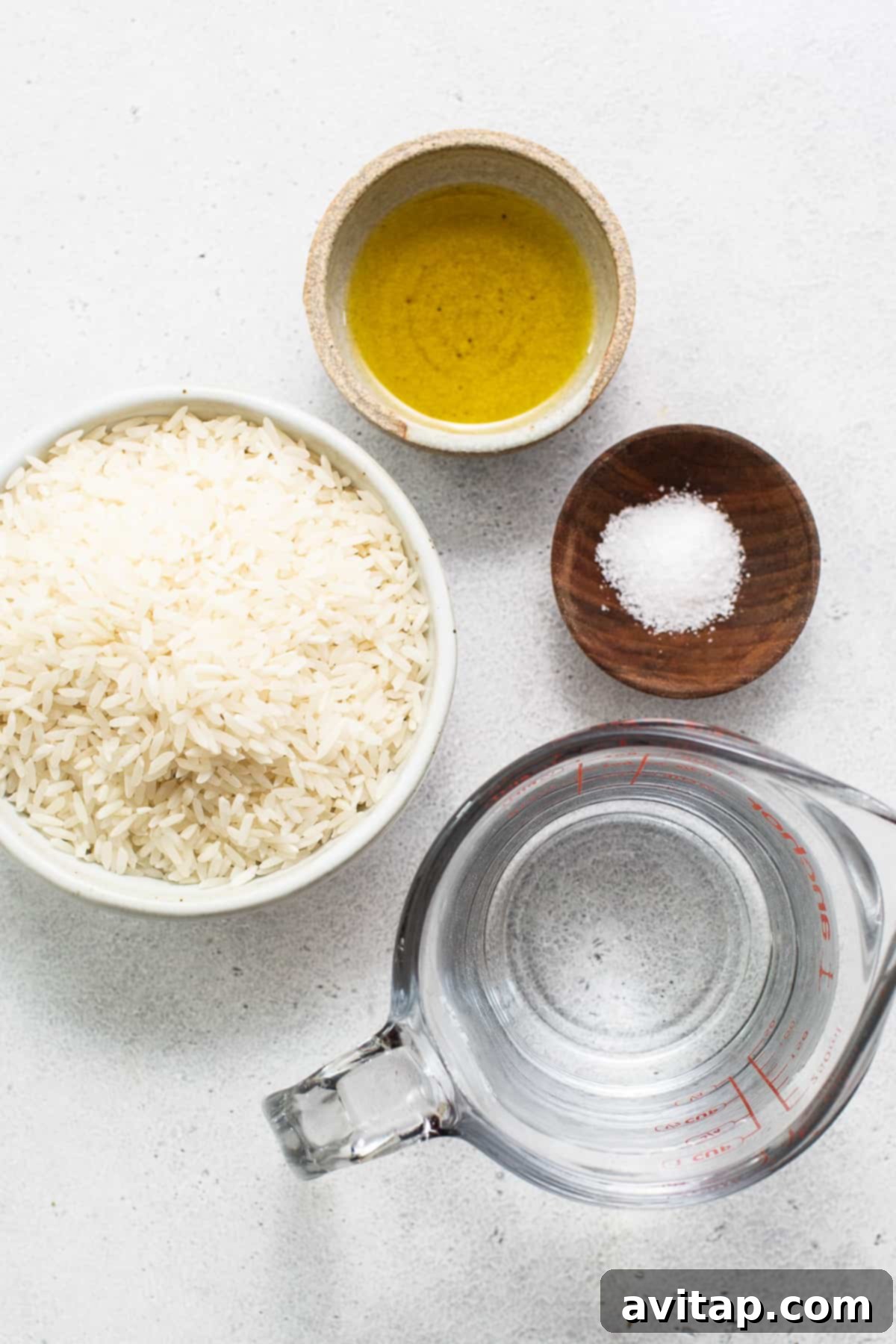 A selection of ingredients for cooking white rice: a bowl of raw rice, a bottle of olive oil, and a small dish of salt, all on a pristine white surface.